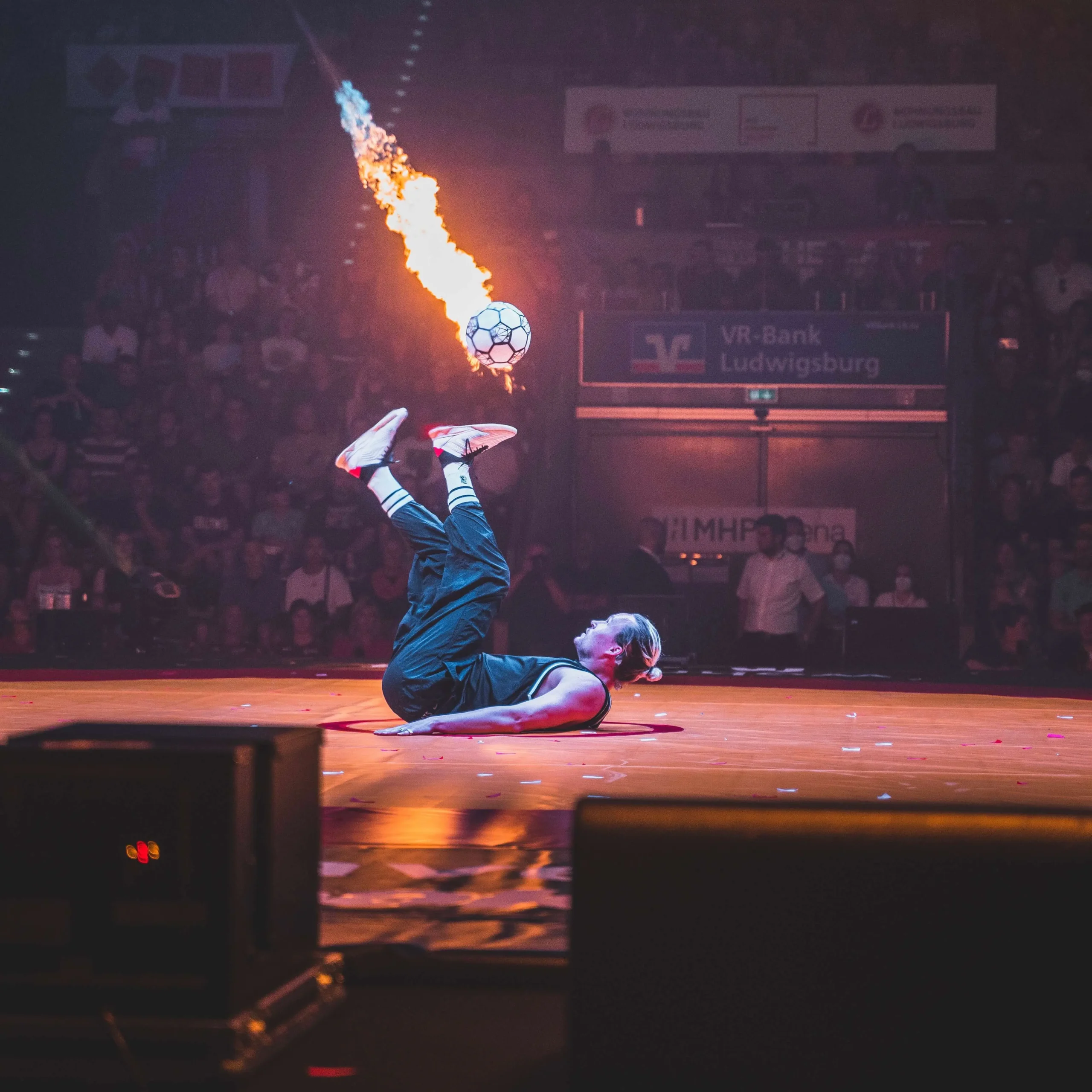 Ein Fußball-Freestyler liegt auf dem Rücken auf der Bühne und schießt einen Fußball in die Luft, aus dem Flammen schießen, während das staunende Publikum im Hintergrund zusieht.