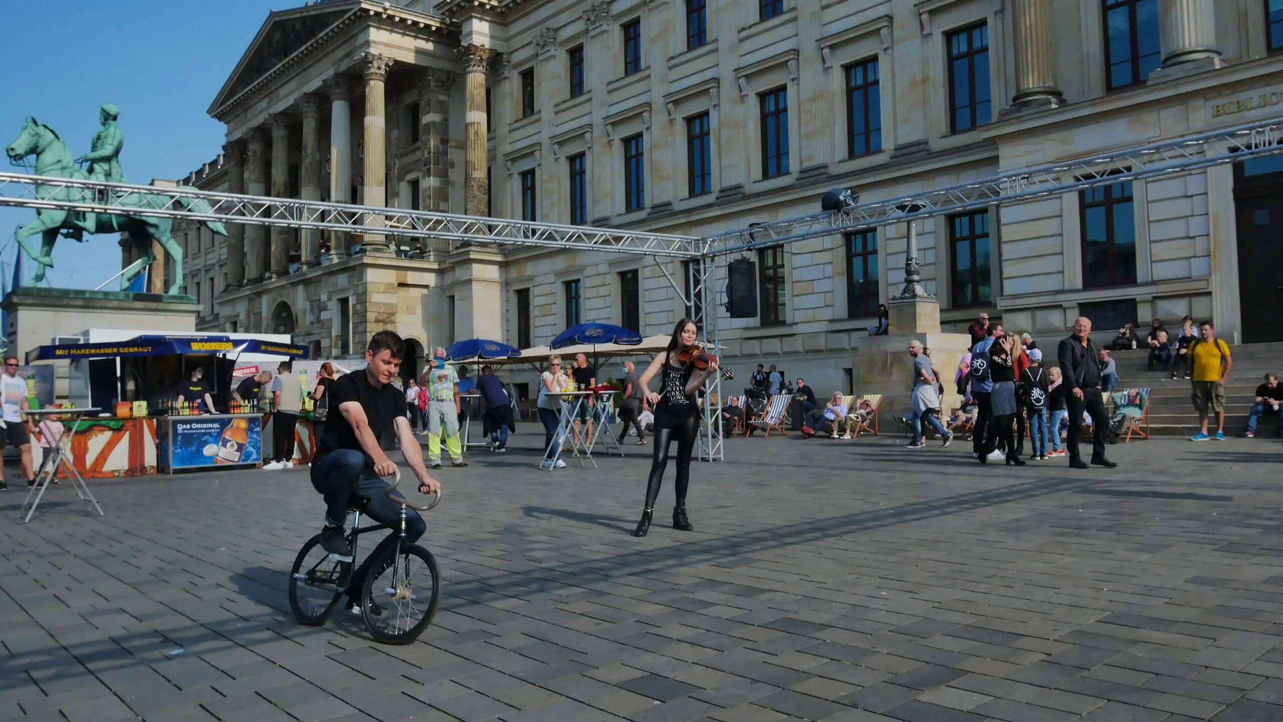Eine Person fährt mit dem Fahrrad über einen belebten Platz, auf dem Menschen spazieren und sitzen. Im Hintergrund sind ein historisches Gebäude, Essensstände und ein Reiterstandbild zu sehen, während eine Kunstrad&Violine-Show den klaren, sonnigen Tag belebt.