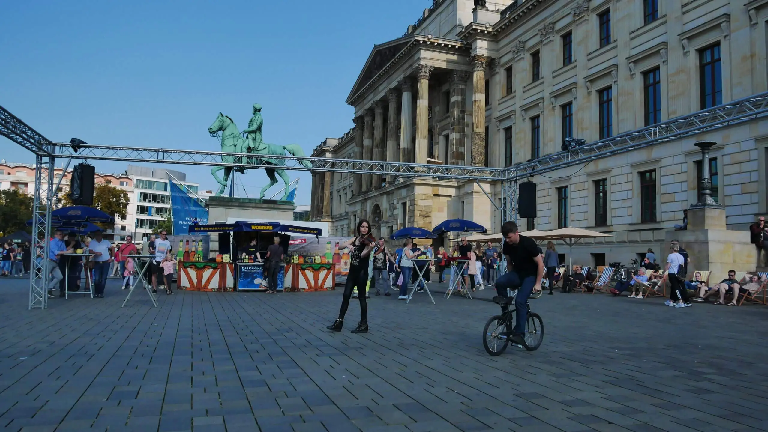 Menschen versammeln sich auf einem Stadtplatz mit Kopfsteinpflaster; ein Mann fährt auf einem BMX-Rad, während eine Kunstrad&Violine-Show in der Nähe einer Frau die Aufmerksamkeit auf sich zieht, während ein Reiterstandbild und historische Gebäude in den klaren Himmel ragen.