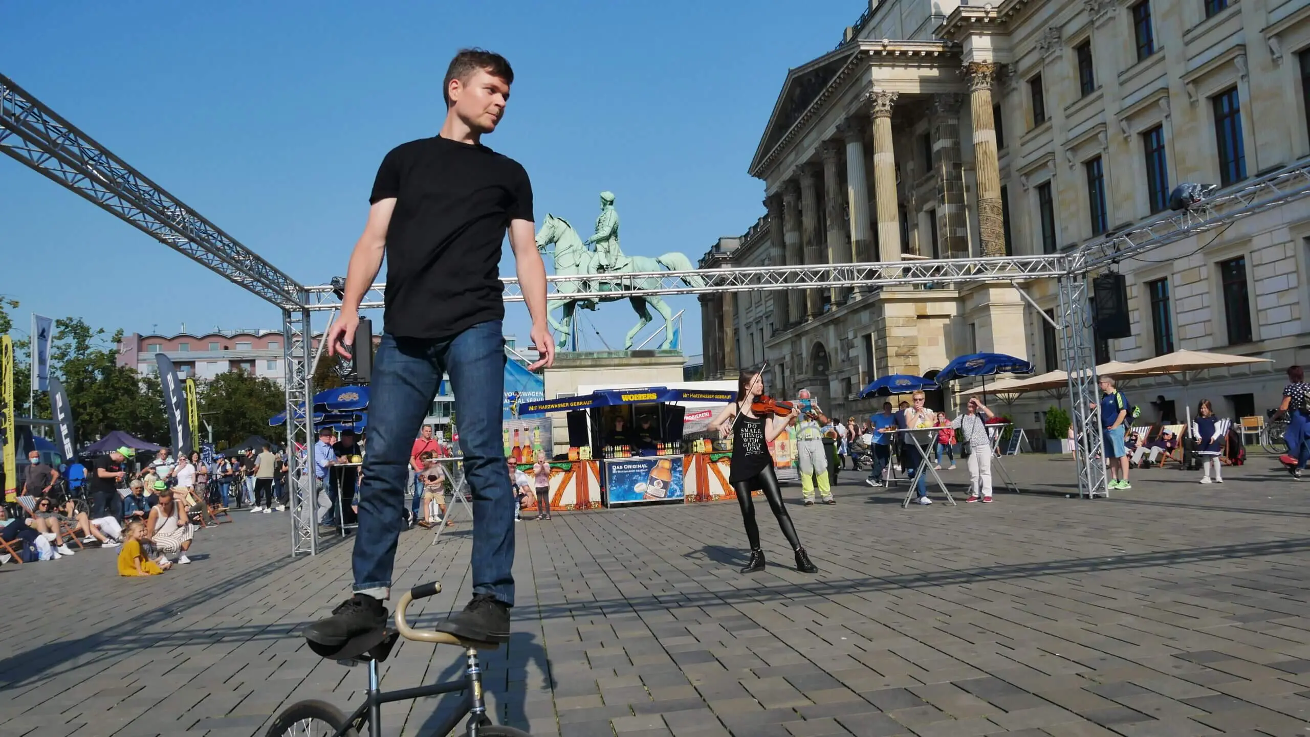 Ein junger Mann in schwarzem T-Shirt und Jeans balanciert auf dem Lenker eines BMX-Rads auf einem belebten Stadtplatz und führt eine Kunstrad&Violine-Show inmitten von Menschen, einer Statue eines Pferdes und eines Reiters sowie historischen Gebäuden im Hintergrund auf.