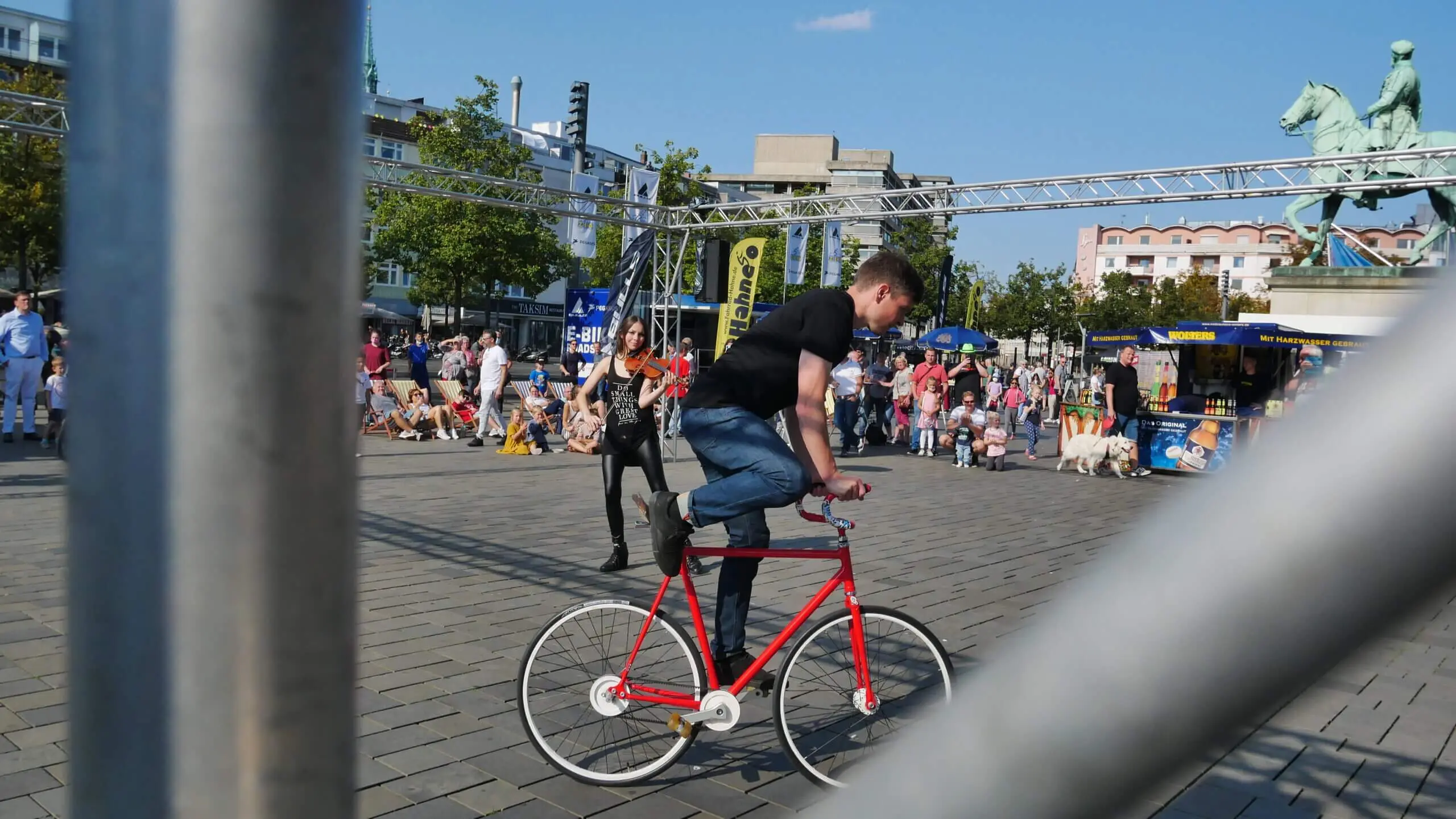 Ein Mann in schwarzem Hemd und Jeans fährt auf einem roten Fahrrad auf einem belebten Stadtplatz, während die Menschen zusehen und der Szene das Flair einer Kunstrad&Violine-Show verleihen. Der Vordergrund ist teilweise durch einen grauen Metallzaun verdeckt.