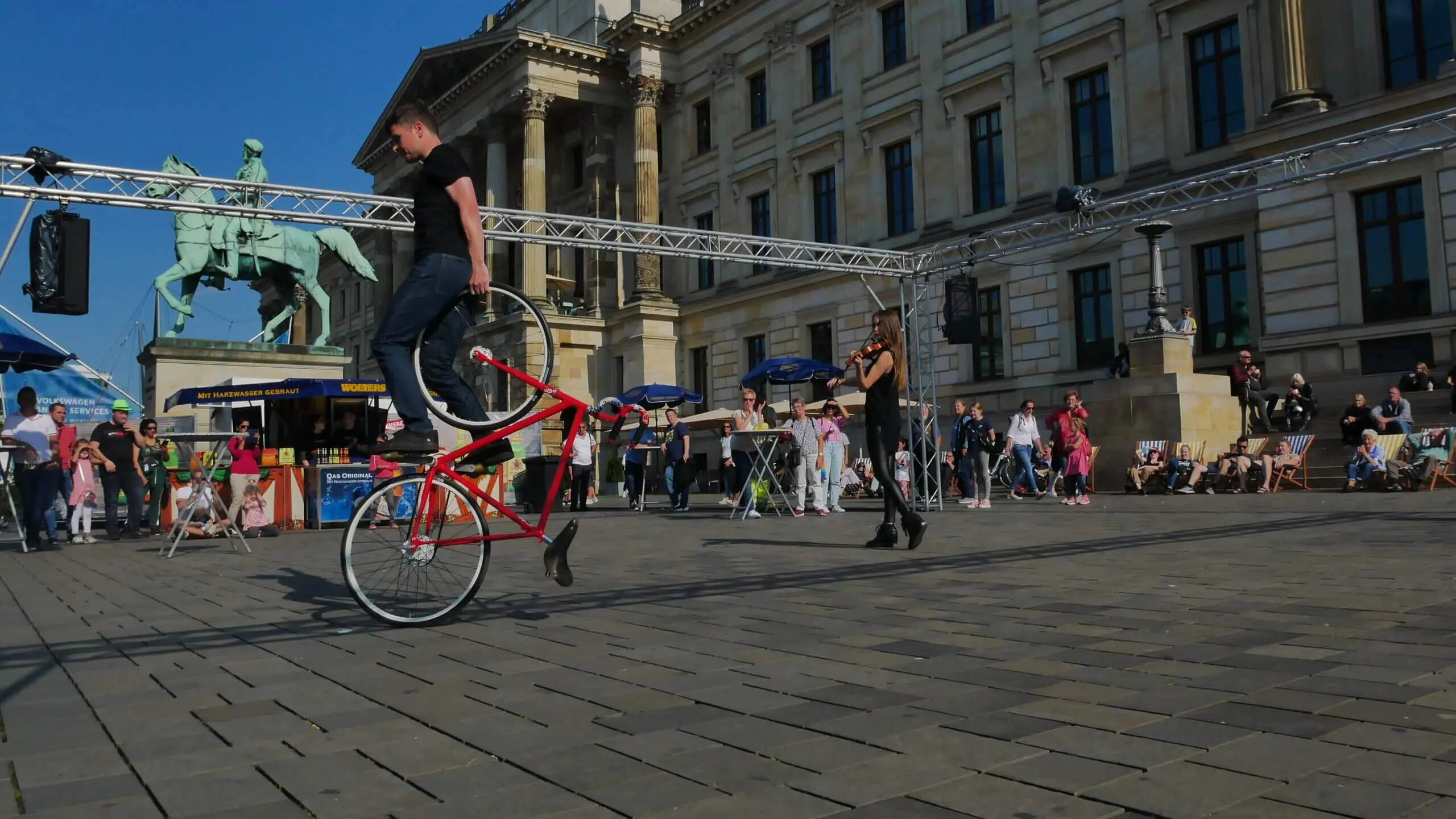 Ein Mann balanciert auf dem Vorderrad eines roten Fahrrads auf einem belebten städtischen Platz während einer Kunstrad&Violine-Show, während im Hintergrund Menschen und historische Gebäude zu sehen sind.