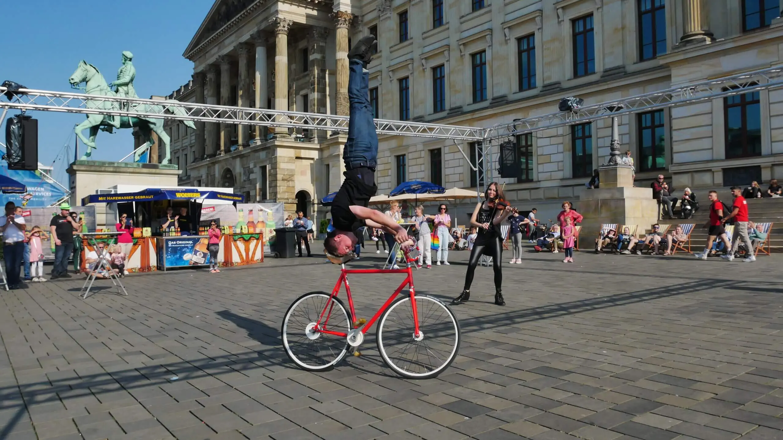Ein Mann macht einen Handstand auf dem Lenker eines roten Fahrrads auf einem Stadtplatz während der Kunstrad&Violine Show, während eine Frau ihn fotografiert. Zwischen historischen Gebäuden und belebten Marktständen schauen die Menschen zu.