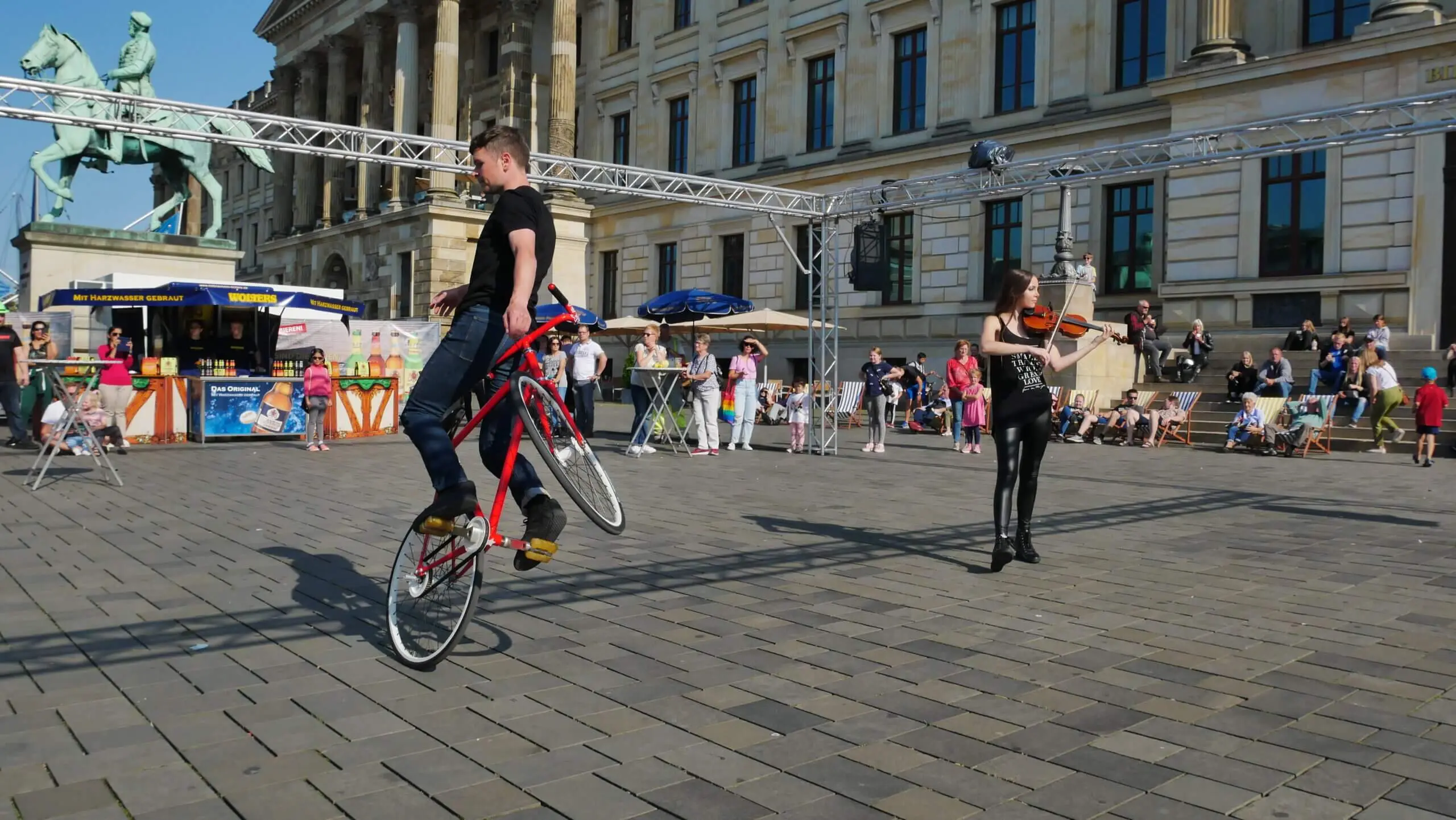 Ein Mann vollführt einen Wheelie auf einem Fahrrad, während eine Frau in der Nähe in einer lebendigen Kunstrad&Violine Show Geige spielt. Sie unterhalten die Menge auf einem Stadtplatz, umgeben von historischen Gebäuden und Schaulustigen.