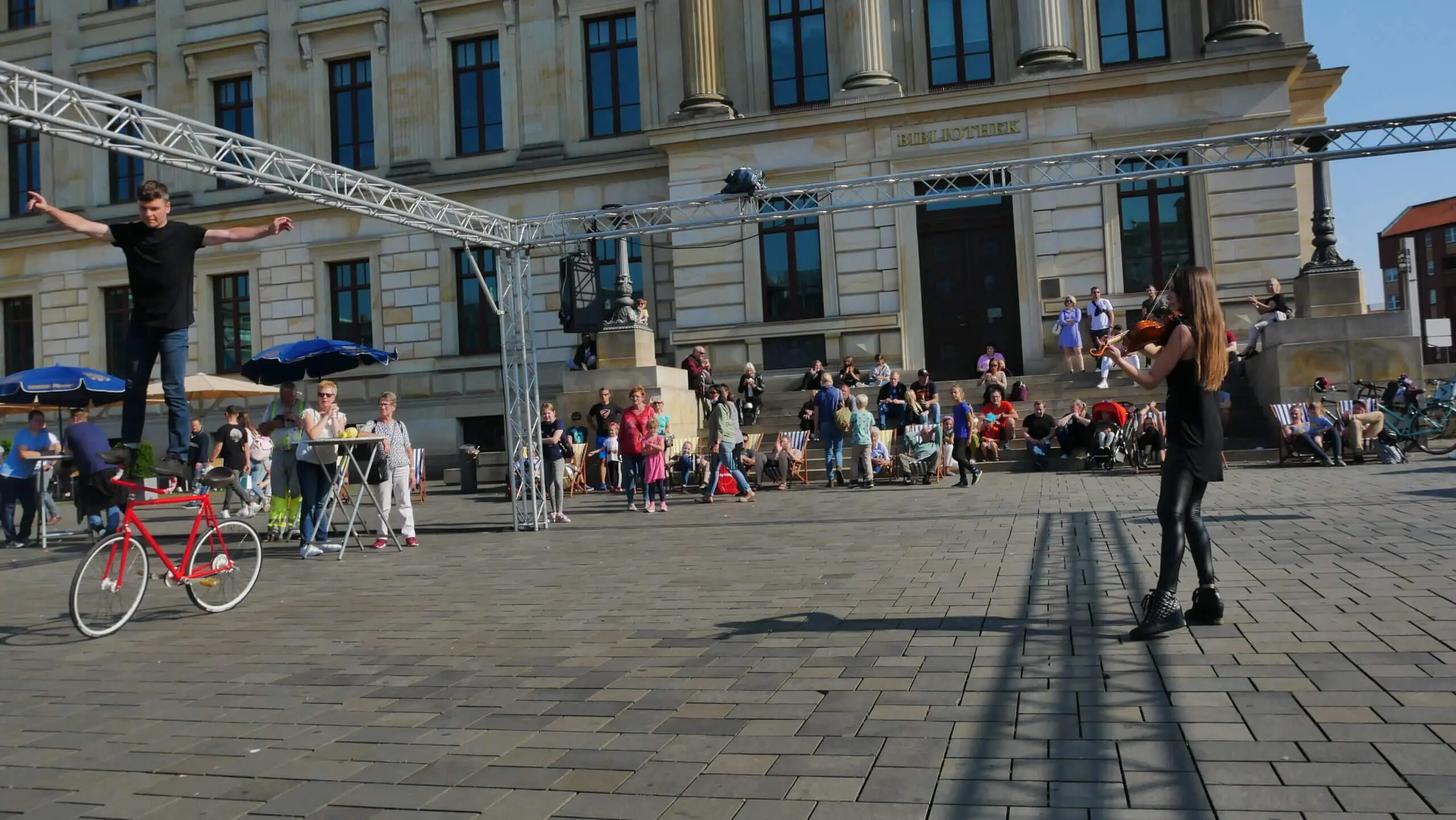 Ein Straßenkünstler der Kunstrad&Violine Show balanciert auf einem Drahtseil über einem Platz und zieht die Zuschauer in seinen Bann. Das historische Gebäude mit seinen Säulen und hohen Fenstern bildet die Kulisse, während in der Nähe ein rotes Fahrrad geparkt ist.