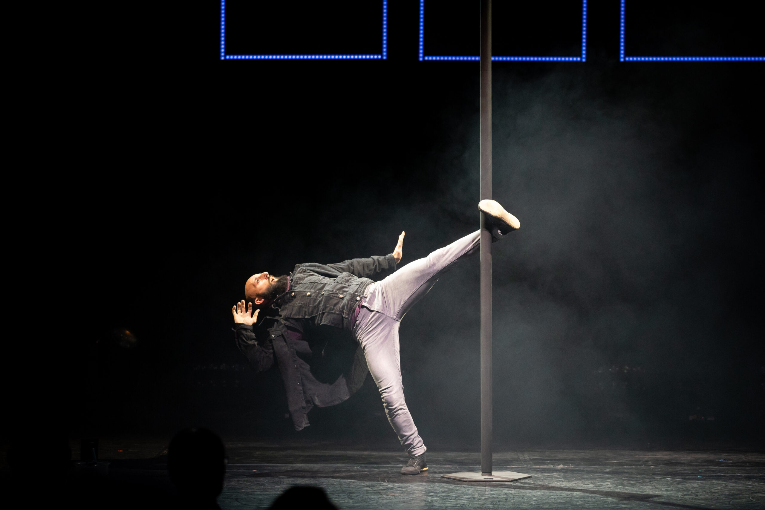 Ein Darsteller in Freizeitkleidung führt einen beeindruckenden High Kick aus, während er eine vertikale Stange auf einer schwach beleuchteten Bühne umgreift, umgeben von dramatischem Rauch und dunklem Hintergrund.
