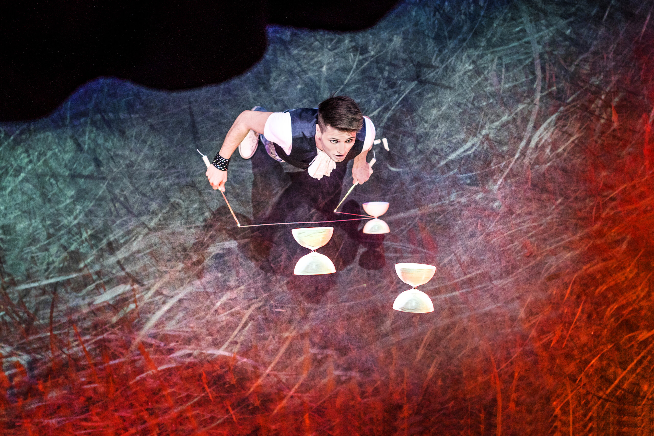 Ein Zirkusartist in einem schwarz-weißen Kostüm jongliert drei weiße Diabolos auf einer bunten, strukturierten Bühne, die von roter und blauer Beleuchtung erhellt wird, von oben gesehen.