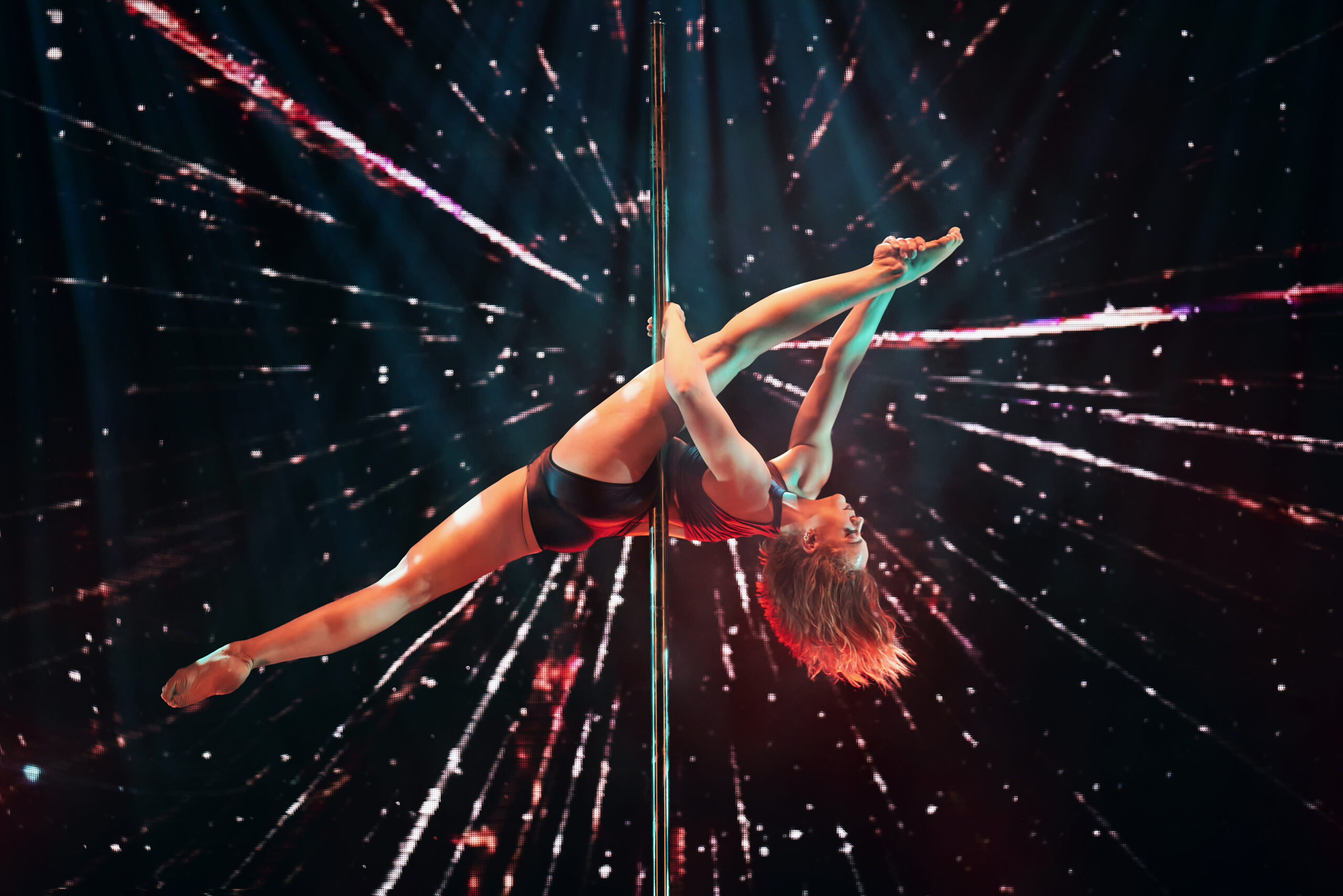 Eine Frau führt eine akrobatische Pose auf einer Stange aus, wobei sie ein Bein nach oben streckt. Sie ist umgeben von einem dramatischen Lichteffekt im Hintergrund, der eine dynamische und energiegeladene Szene erzeugt.