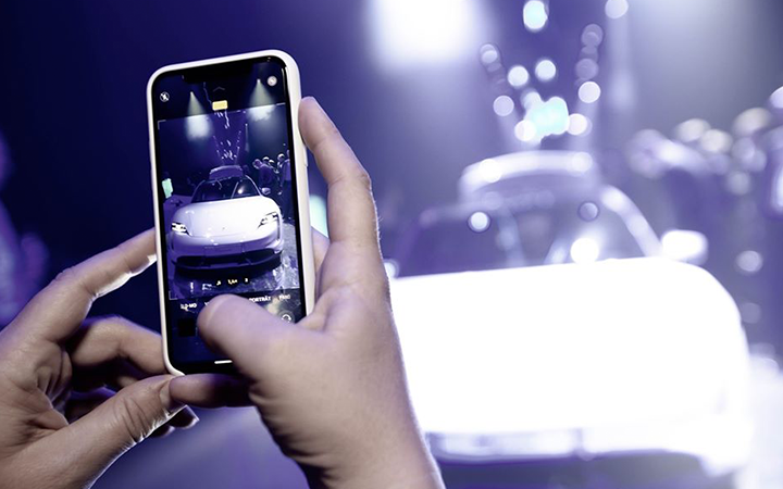 Eine Person hält ein Smartphone in der Hand und fotografiert ein weißes Auto, das auf einer LED-Show ausgestellt ist, mit hellen, verschwommenen Lichtern im Hintergrund. Auf dem Bildschirm des Smartphones ist das Auto deutlich zu erkennen.