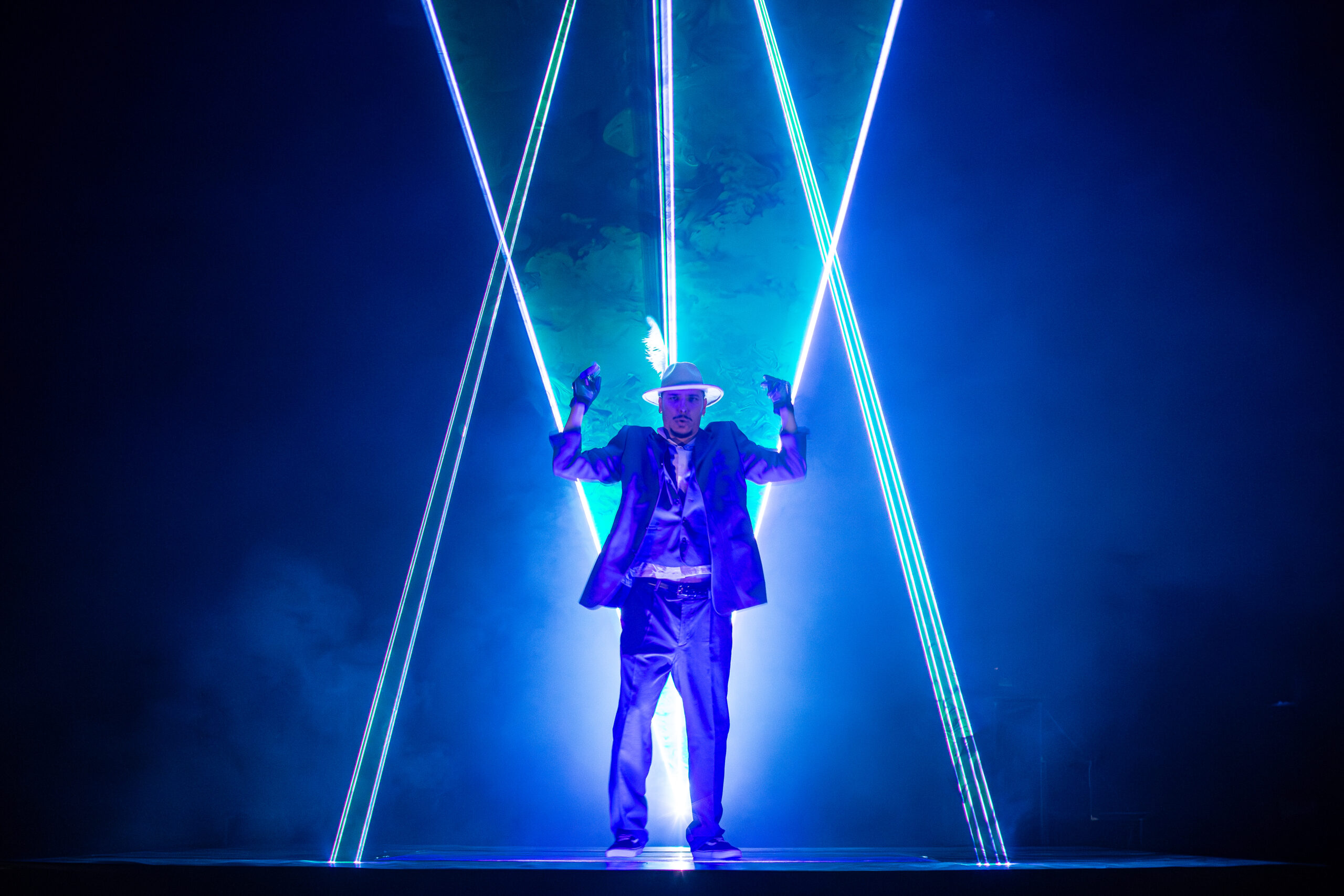 Laser Dance Show .Eine Person in Anzug und Hut steht auf der Bühne, umgeben von sich kreuzenden blauen und weißen Laserstrahlen, mit einem dramatischen blauen Leuchten und Rauch im Hintergrund.