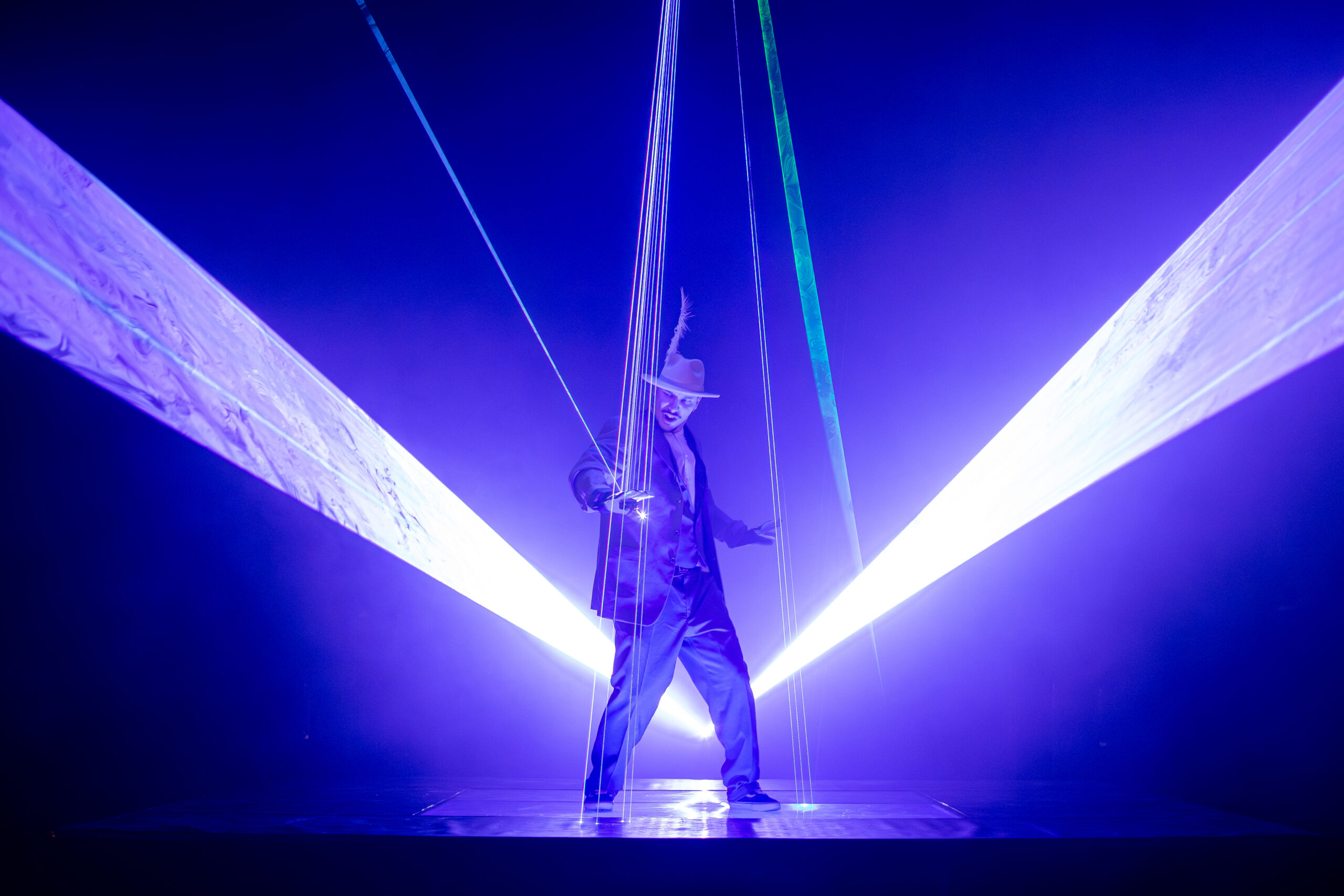 Laser Dance Show .Ein Darsteller in Anzug und Hut steht auf der Bühne inmitten heller, bunter Laserstrahlen und blau-violettem Licht, die eine dramatische und futuristische Atmosphäre schaffen.