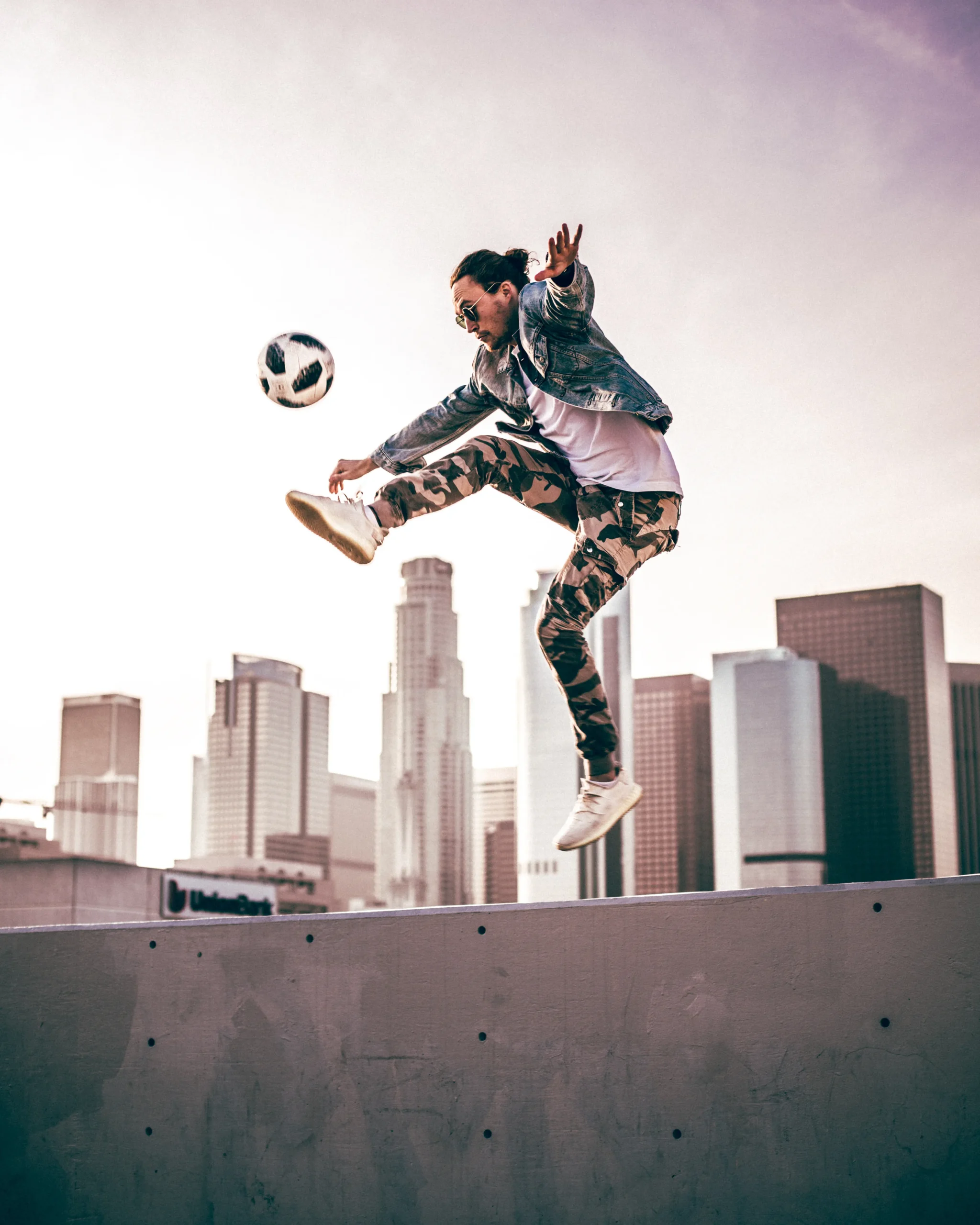 Ein Fußball-Freestyler in Jeansjacke und Camouflage-Hose springt in die Luft, während er einen Fußball kickt, mit hohen Stadtgebäuden und einem rosafarbenen Himmel im Hintergrund.