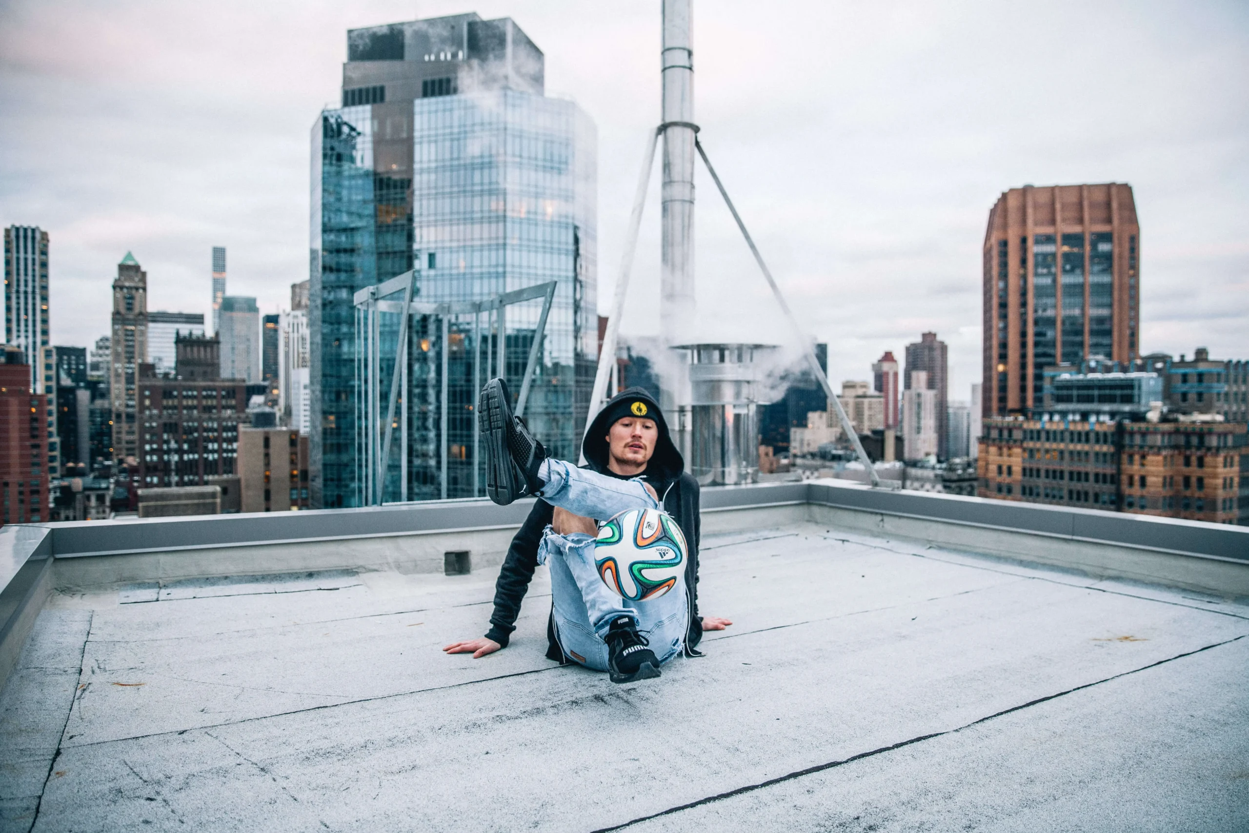 Ein Fußball-Freestyler sitzt mit ausgestreckten Beinen auf einem Dach und balanciert gekonnt einen Fußball auf einem Fuß. Im Hintergrund ragen Wolkenkratzer und Gebäude unter einem wolkenverhangenen Himmel in die Höhe.