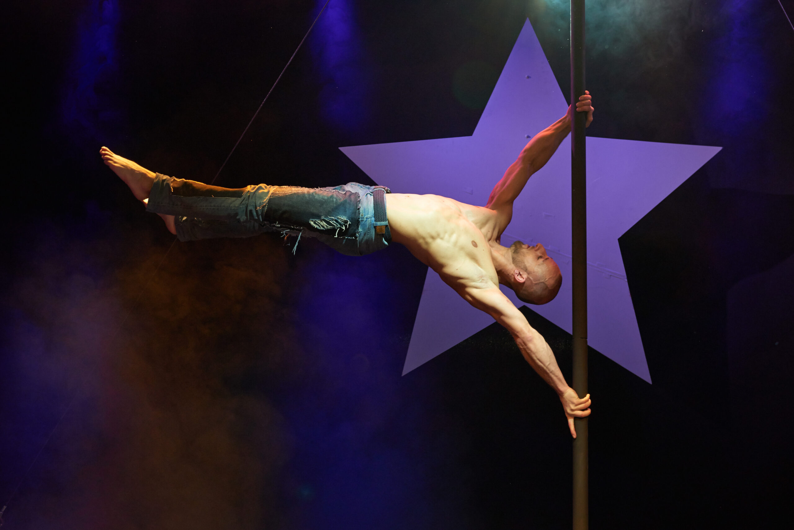 Ein hemdsärmeliger Mann in zerrissenen Jeans führt auf der Bühne einen horizontalen Griff an einer vertikalen Stange aus, während im Hintergrund ein großer weißer Stern und bunte Bühnenlichter zu sehen sind.