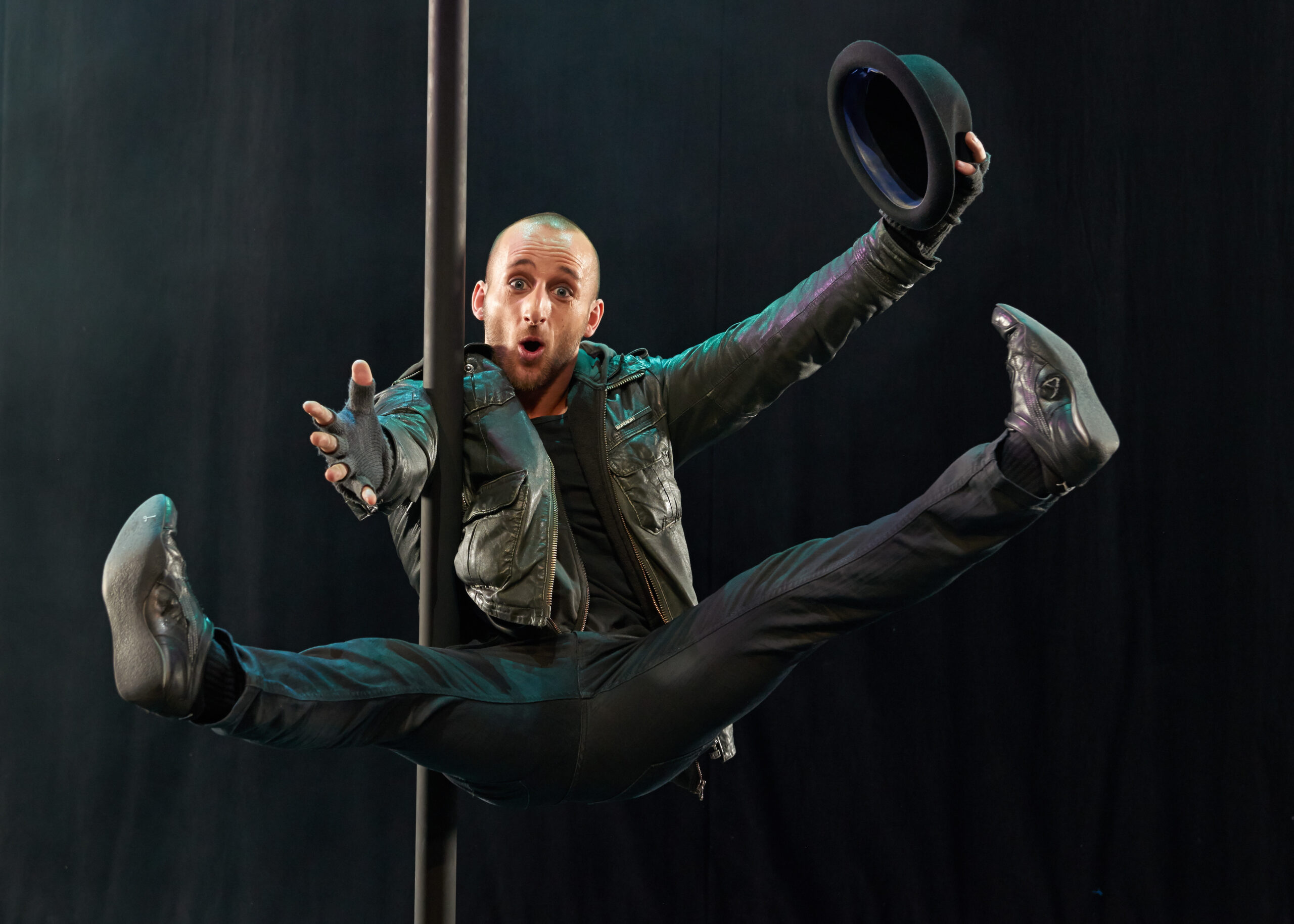 Ein Mann mit Lederjacke, Handschuhen und Hut führt einen Spagatsprung aus, während er eine vertikale Stange umgreift, mit einem überraschten Gesichtsausdruck vor einem dunklen Hintergrund.
