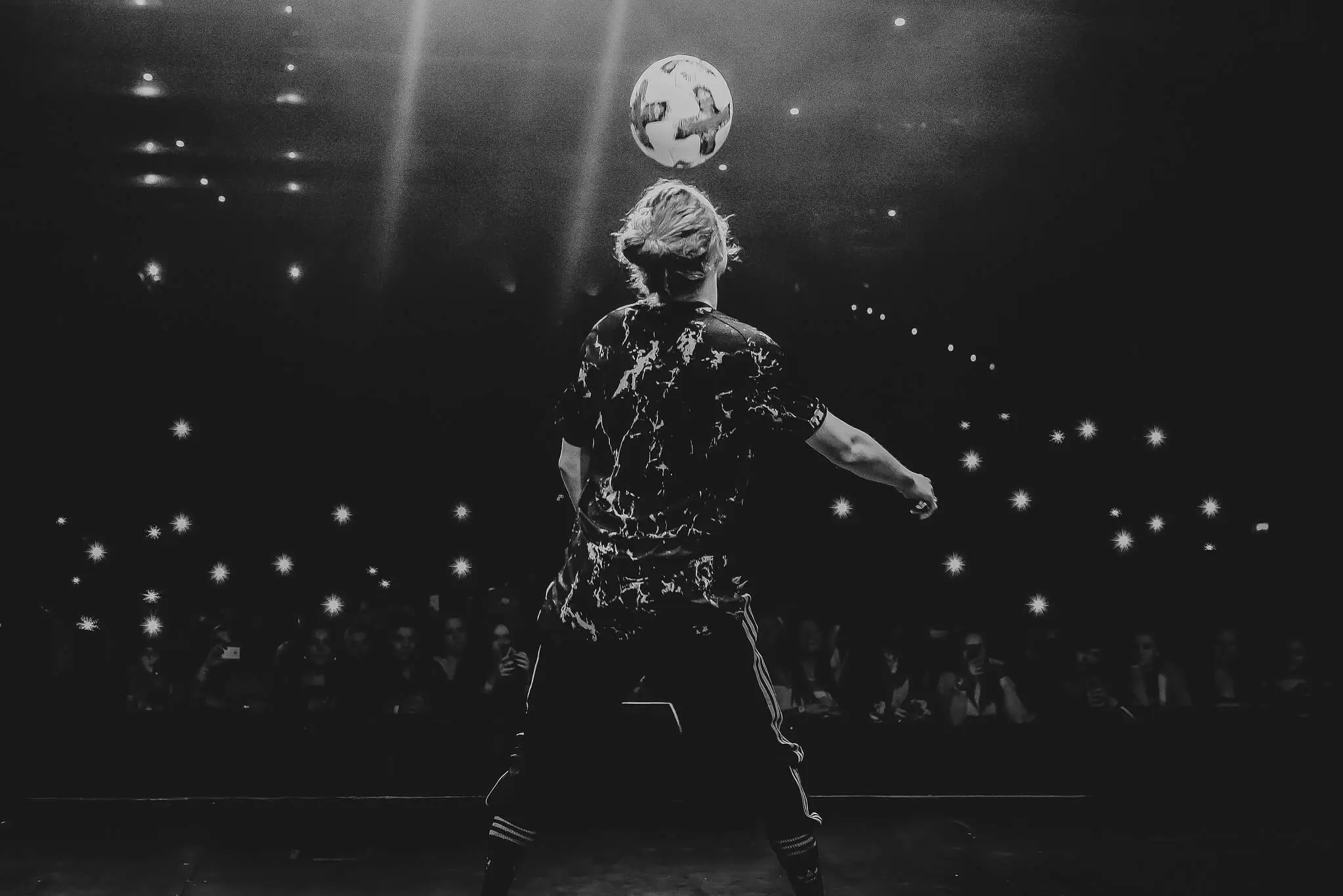 Ein Fußball-Freestyler in Sportkleidung steht auf der Bühne vor einem Publikum und balanciert einen Fußball auf seinem Kopf. Die Zuschauer halten leuchtende Handys in die Höhe und erzeugen Lichtpunkte im dunklen Hintergrund.