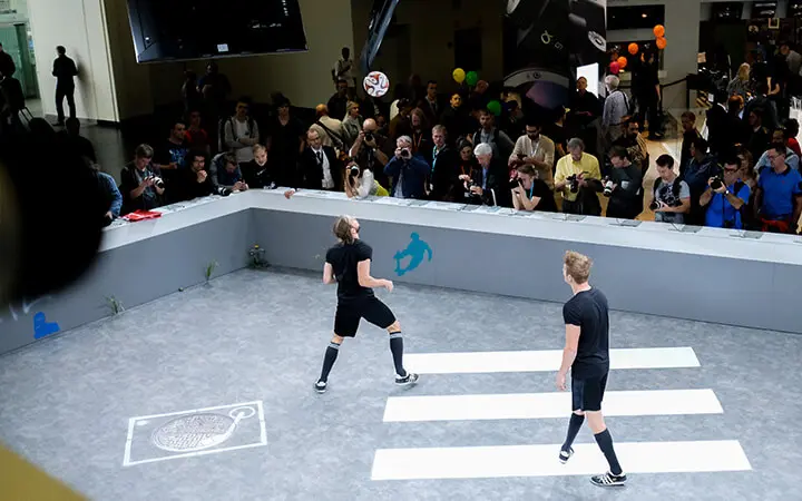Zwei Personen in schwarzer Sportkleidung spielen während aufregender Messeshows Fußball auf einem kleinen Hallenplatz, während eine Menschenmenge zusieht. Ein Spieler schießt den Ball in die Luft; Linien und Grafiken zieren den grauen Boden, während das Publikum hinter Absperrungen steht und das Spiel beobachtet.