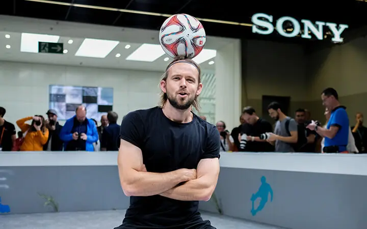 Ein Mann in einem schwarzen Hemd balanciert während einer beeindruckenden Messeshow mit verschränkten Armen einen Fußball auf dem Kopf, während im Hintergrund Menschen Fotos machen. Über ihm ist das SONY-Logo zu sehen.