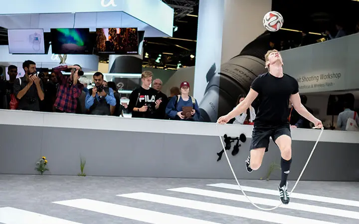 Ein Mann in Sportkleidung springt Seil, während er einen Fußball auf der Stirn balanciert. Er begeistert die Menge mit seiner Messeshow-Performance, während die Zuschauer den Moment mit ihren Handys festhalten.