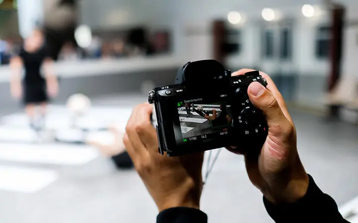 Eine Person hält auf einer Messeshow-Veranstaltung eine Digitalkamera in der Hand und konzentriert sich auf eine Szene mit einer unscharfen Person im Hintergrund. Der Bildschirm der Kamera zeigt ein klareres Bild der Szene, die in einem Innenraum fotografiert wird.