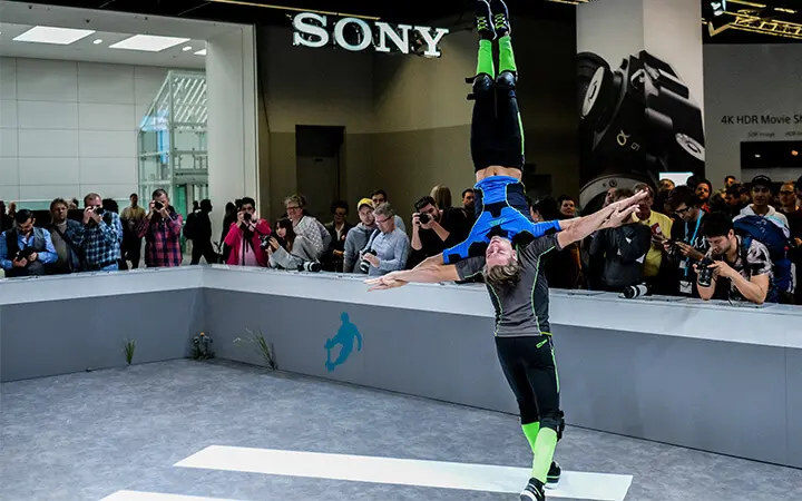 Zwei Akrobaten vollführen einen Balanceakt; einer hält den anderen kopfüber an der Taille. Sie begeistern das Publikum mit ihrer Messeshow auf einer Technikmesse, wobei im Hintergrund ein großes SONY-Schild und Kameras zu sehen sind.
