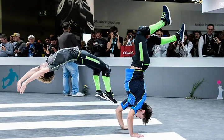 Zwei Artisten in grün-schwarzen Anzügen verblüffen das Publikum mit beeindruckenden Messeshows, bei denen der eine einen Rückwärtssalto vollführt und der andere auf den Händen balanciert, während das Publikum im Hintergrund Fotos schießt.