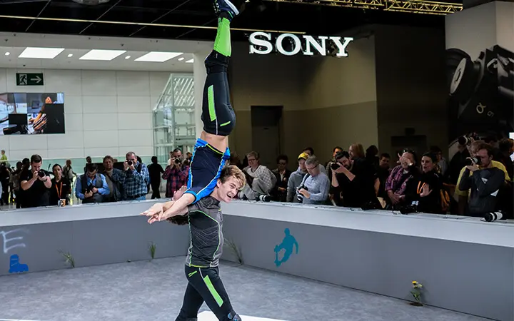 Zwei Akrobaten vollführen bei den Messeshows einen Balanceakt; einer steht aufrecht und hält den anderen kopfüber an der Taille. Ein Publikum schaut zu und macht Fotos, im Hintergrund ist ein großes "SONY"-Schild zu sehen.