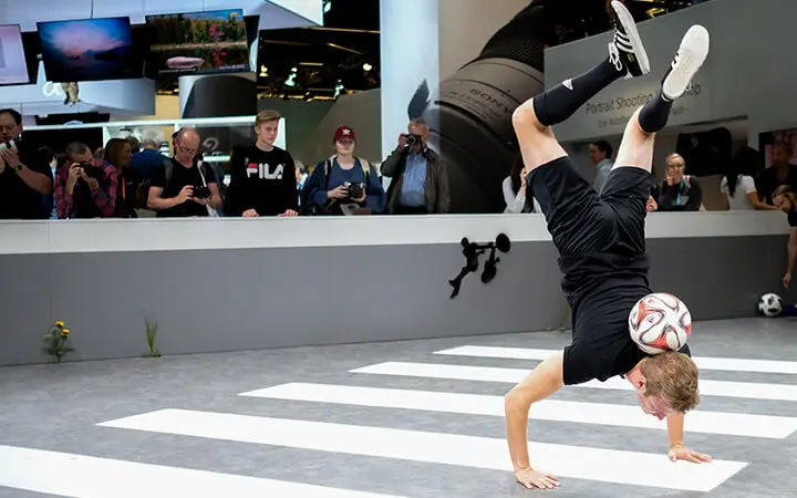 Eine Person balanciert auf einer Hand, während sie einen Fußball zwischen Kopf und Arm hält, während einer beeindruckenden Messeshow-Performance auf einem gestreiften Boden, während eine Menschenmenge im Hintergrund zusieht und Fotos macht.