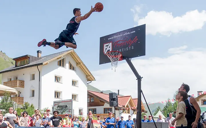 Bei der Basketball Dunking Show fliegt ein Basketballspieler durch die Luft und erzielt einen Slam Dunk, während die Zuschauer zusehen und die Gebäude im europäischen Stil eine atemberaubende Kulisse bilden.