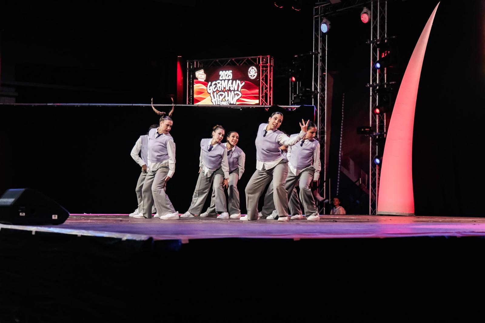 Eine Streetdance-Tanzgruppe in passenden grauen Outfits tritt auf der Bühne unter bunten Lichtern bei der Veranstaltung 2023 Germany Hip Hop International auf. Ein großes digitales Schild zeigt den Namen der Veranstaltung im Hintergrund an.
