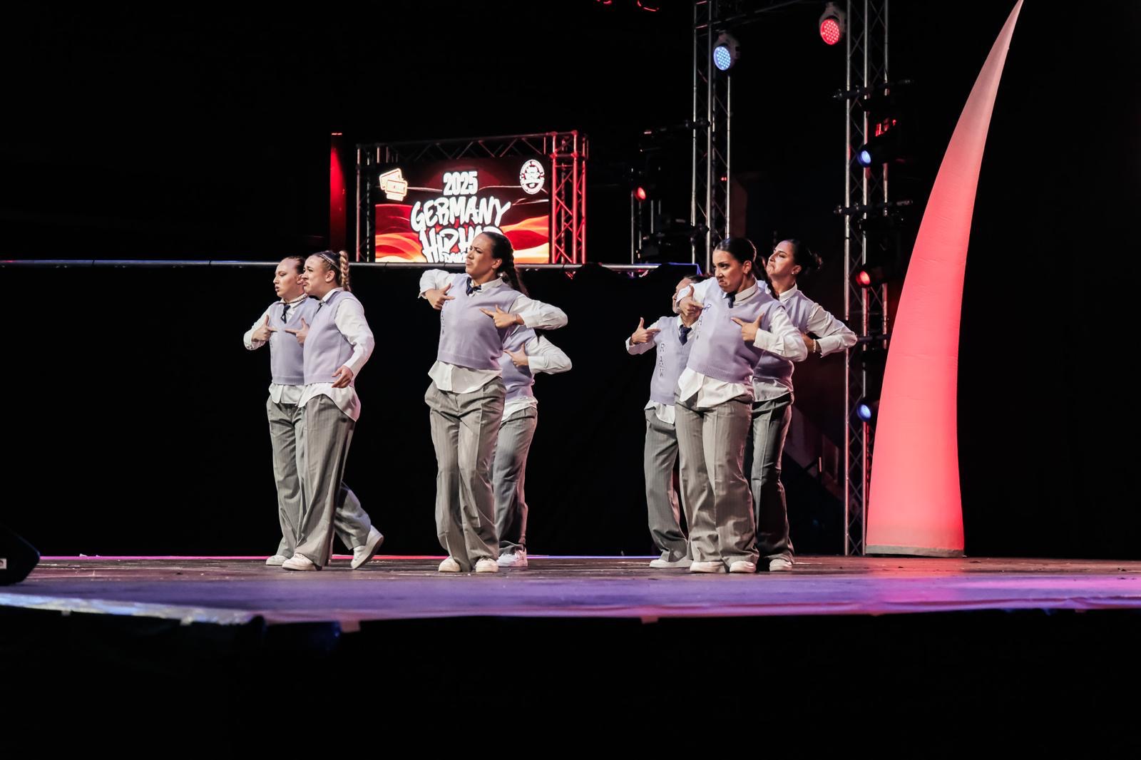 Eine Gruppe von Tänzerinnen und Tänzern in passenden grau-weißen Outfits führt auf der Bühne des Hip-Hop-Events 2023 Deutschland eine synchrone Nummer auf, mit bunten Lichtern und einem Schild im Hintergrund.