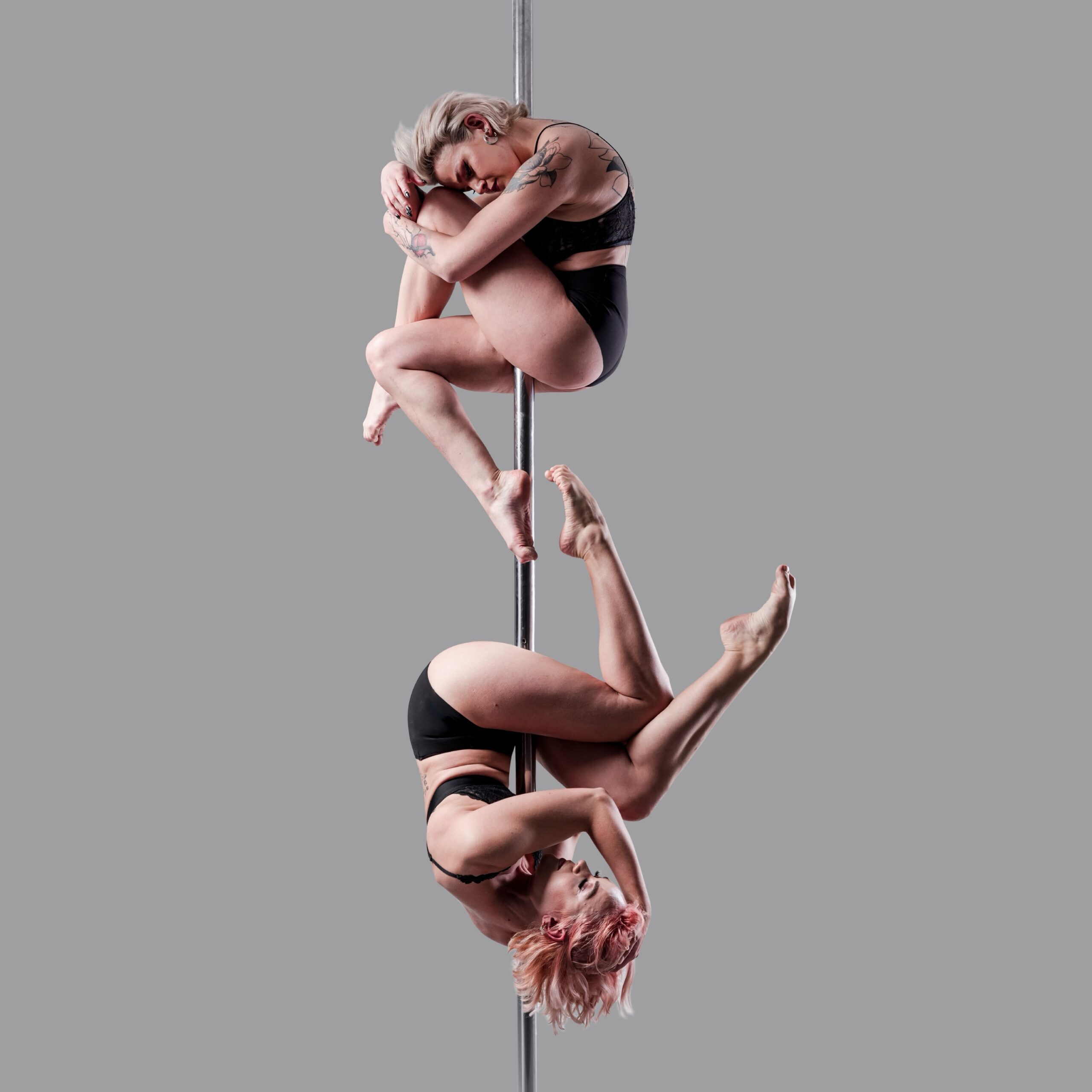 Zwei Frauen in schwarzer Sportkleidung führen fortgeschrittene Posen an einer vertikalen Stange aus, wobei jede eine andere Pose einnimmt, vor einem neutralen grauen Hintergrund.