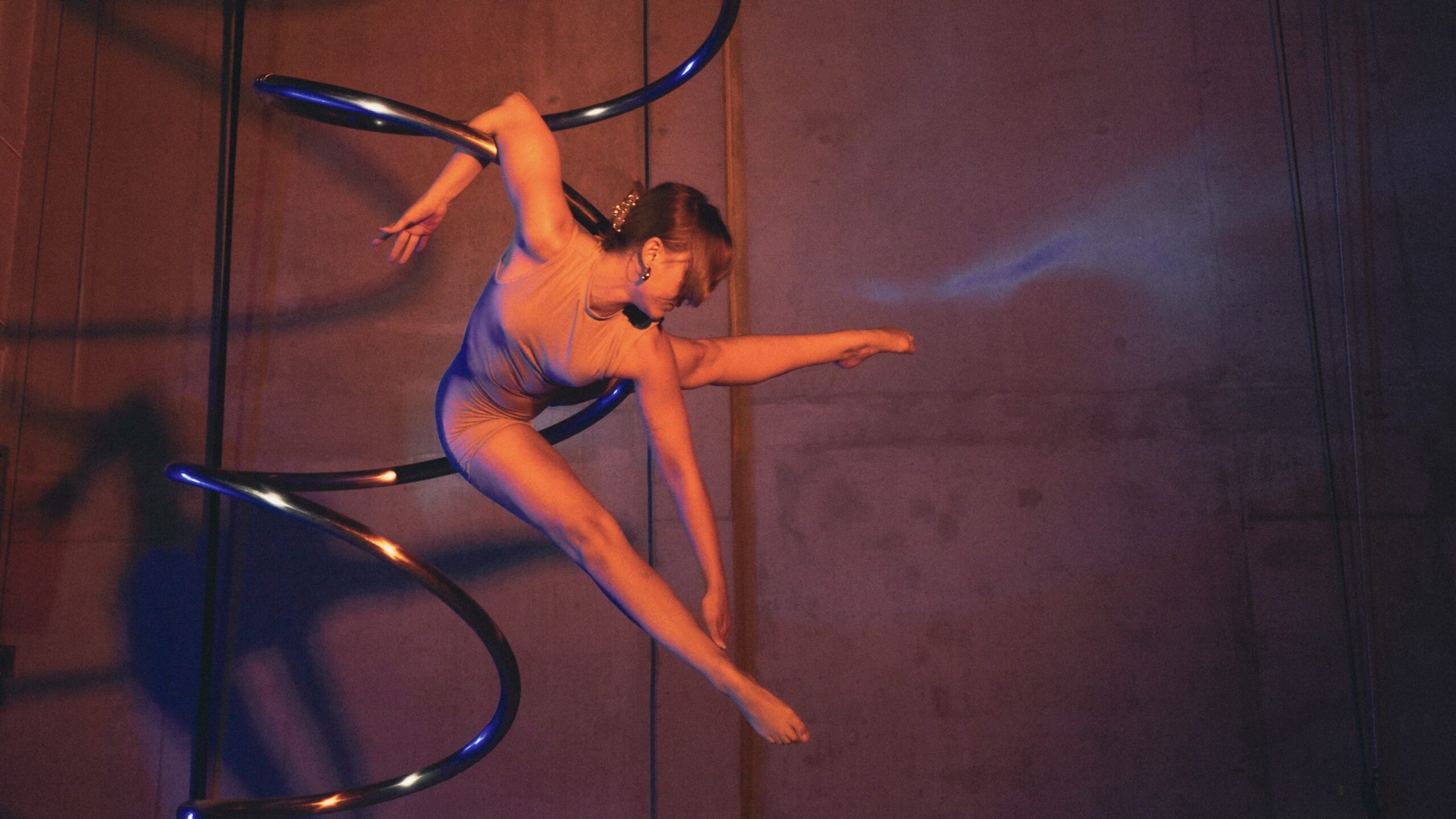 Eine Darstellerin in einem beigen Trikot posiert anmutig an einem spiralförmigen Luftreifen, während eine dramatische Beleuchtung Schatten auf eine schlichte Wand hinter ihr wirft.