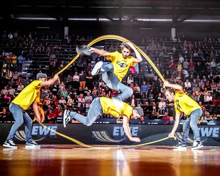 Vier Personen in gelben Hemden führen während einer Halbzeit-Show auf dem Boden einer Turnhalle eine doppelte holländische Seilsprungübung vor, bei der eine Person hoch springt, während zwei Personen Seile drehen und eine weitere Person niedrig springt; ein großes Publikum schaut im Hintergrund zu.