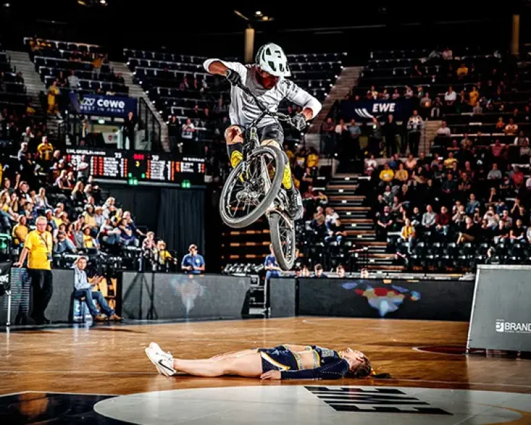 Ein Radfahrer mit Helm springt während einer Hallen-Halbzeit-Show über eine auf einem Basketballfeld liegende Person, während die Zuschauer von der Tribüne aus zusehen.