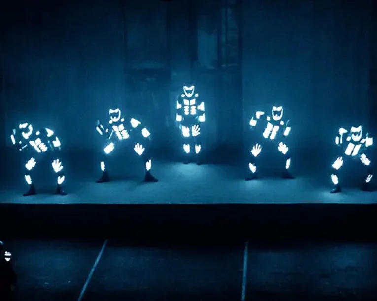 Fünf Künstler in leuchtenden Anzügen mit beleuchteten Mustern stehen auf einer dunklen Bühne und bieten eine beeindruckende LED-Show. Ihre Anzüge weisen leuchtende Formen auf der Brust, den Armen und Beinen auf und sorgen so für eine faszinierende visuelle Darstellung.