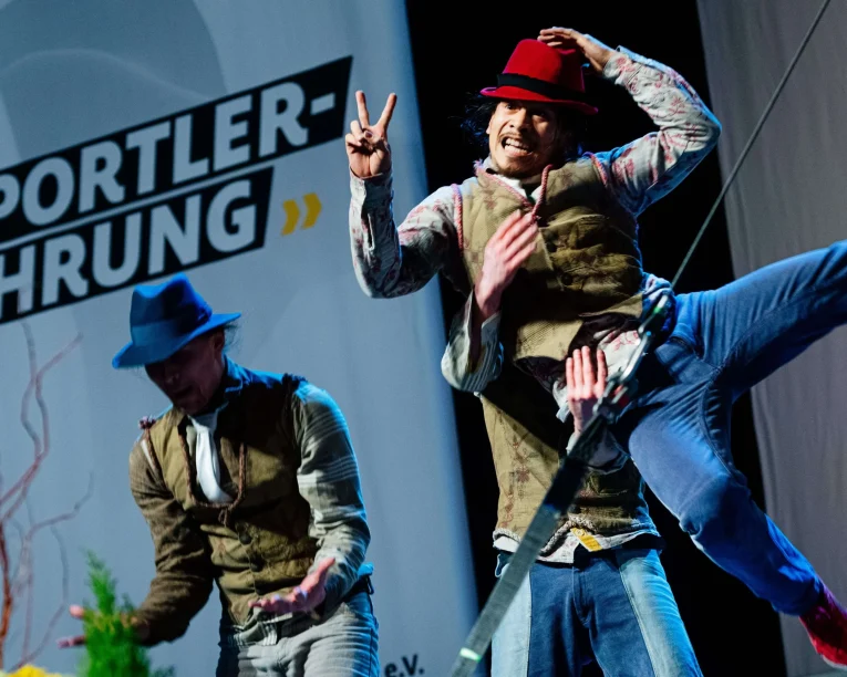 Zwei Darsteller in Westen und Hüten zeigen auf der Bühne eindrucksvolle Comedy-Artistik; einer springt in der Luft und zeigt ein Friedenszeichen. Auf einem großen Schild im Hintergrund steht SPORTLER-EHRUNG.