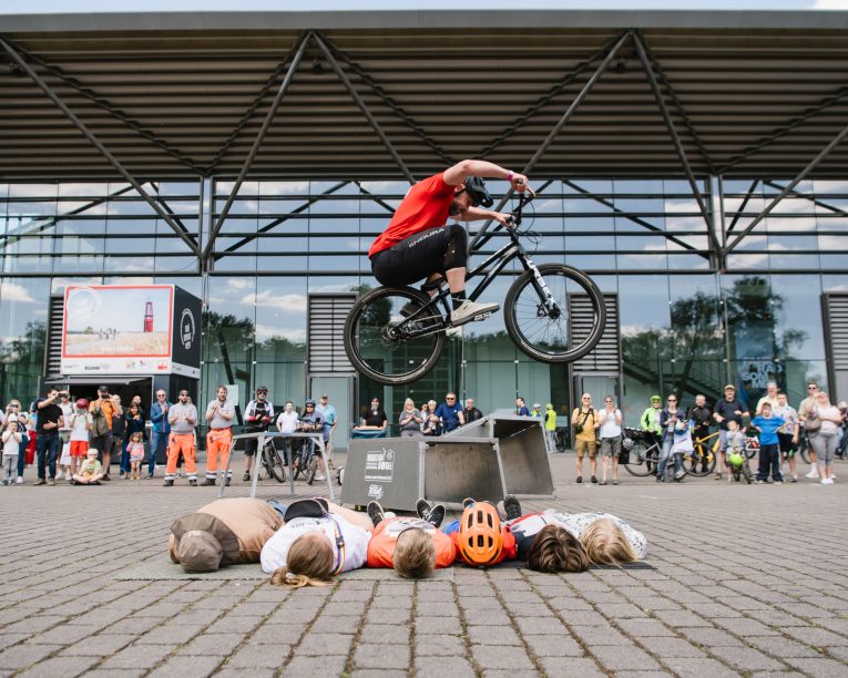 Ein Radfahrer springt mit einem BMX-Rad in der Luft über vier Personen, die nebeneinander auf dem Boden liegen, während eine Menge von Zuschauern vor einem modernen Glasgebäude zusieht.