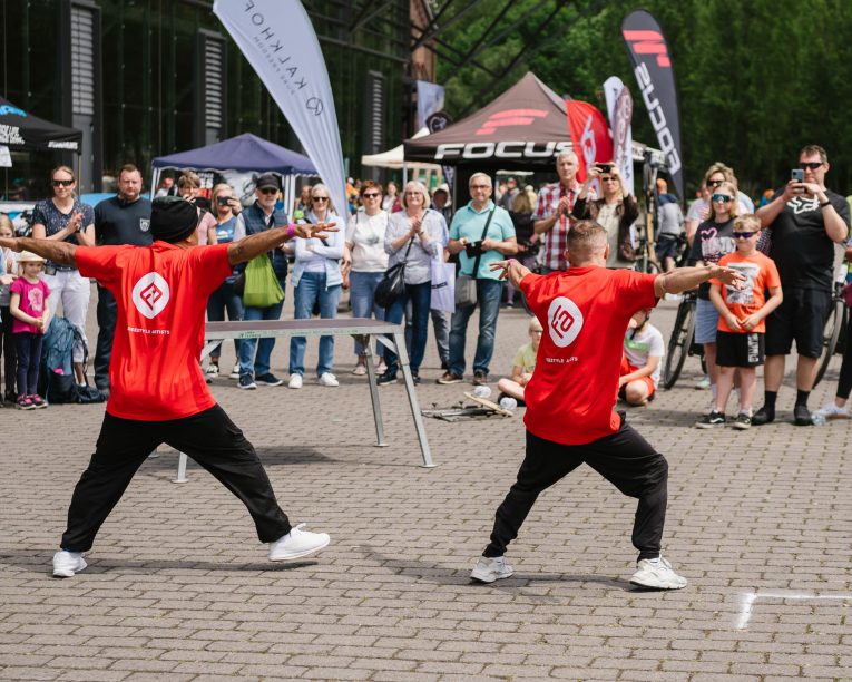 Zwei Darsteller in passenden roten Hemden und schwarzen Hosen tanzen energisch vor einer Menschenmenge im Freien, während Schaulustige in der Nähe von Veranstaltungszelten und Bannern die Aufführung beobachten und aufnehmen.