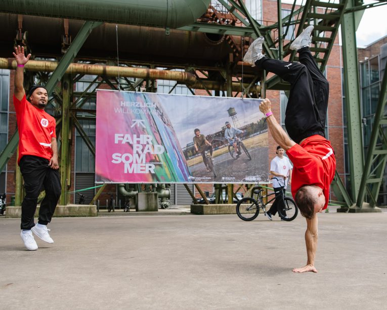 Urban Street Show, Zwei Männer in roten Hemden tanzen Breakdance im Freien unter einer Metallkonstruktion; einer macht einen Handstand, während der andere zusieht. Im Hintergrund sind ein Fahrrad und ein buntes Veranstaltungsbanner zu sehen.