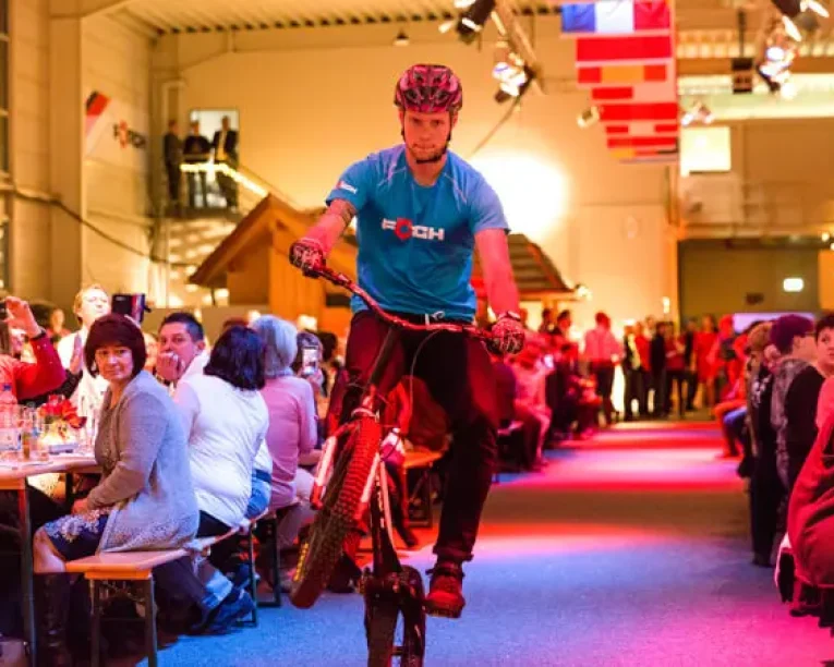 Ein Mann mit Helm und blauem Hemd vollführt einen Wheelie auf einem Fahrrad in einem Innenraum und fährt zwischen langen Tischen auf einer lebhaften Weihnachtsfeier, während die Leute zusehen und Fotos inmitten von festlicher Dekoration und Fahnen machen.