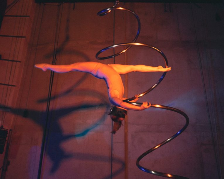 Ein Luftakrobat vollführt einen Spagat, während er an einem großen, spiralförmigen Metallgerät hängt, das auf einer Bühne dramatisch beleuchtet wird.