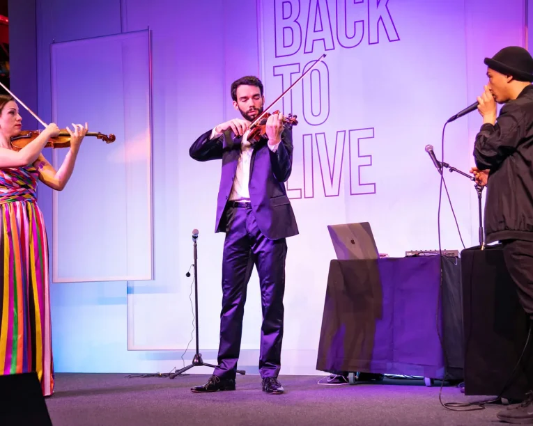 Drei Musiker stehen auf der Bühne; zwei spielen Geige - eine Frau in einem bunten Kleid, ein Mann im Anzug - während ein Mann in dunkler Kleidung und mit Mütze in ein Mikrofon singt. Die Kulisse zeigt Back2Live hinter ihnen.