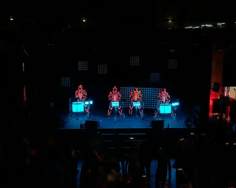 Fünf Künstler in leuchtenden Anzügen begeistern das Publikum mit einer hypnotisierenden LED-Trommel-Show, bei der sie auf einer dunklen Bühne mit blauen Lichtern Schlagzeug spielen. Auf dem überwiegend schwarzen Hintergrund ist rechts im Bild ein beleuchteter Eingang zu sehen.