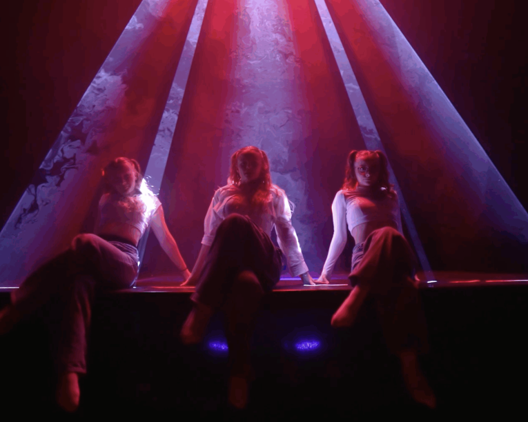 Drei Personen sitzen auf einer dunklen Bühne unter dramatischen roten und violetten Scheinwerfern, die hinter ihnen dreieckige Balken bilden und eine stimmungsvolle, theatralische Atmosphäre schaffen.