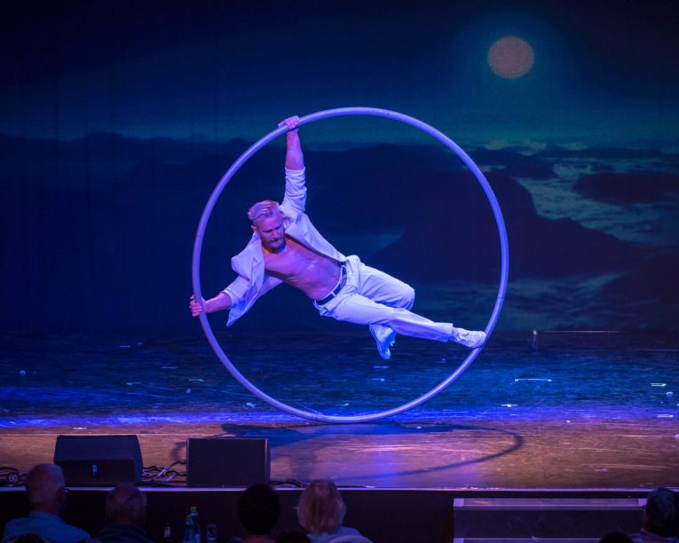 Ein Darsteller in weißer Kleidung balanciert auf einem großen Cyr-Rad auf der Bühne, das sich gerade dreht, vor einer mondbeschienenen Kulisse, während das Publikum im Vordergrund zusieht.