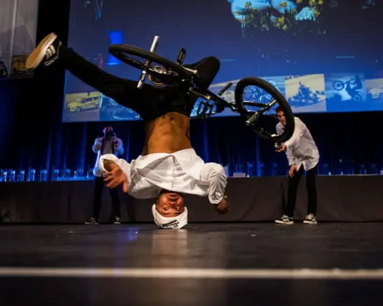 Ein BMX-Fahrer führt einen Kopfstandtrick aus, während er sein Fahrrad in einer Halle hält, wobei er seine Beine und sein Fahrrad während einer spannenden Mix-Show über sich selbst hebt. Zwei Personen schauen im Hintergrund bei blauer Bühnenbeleuchtung zu.