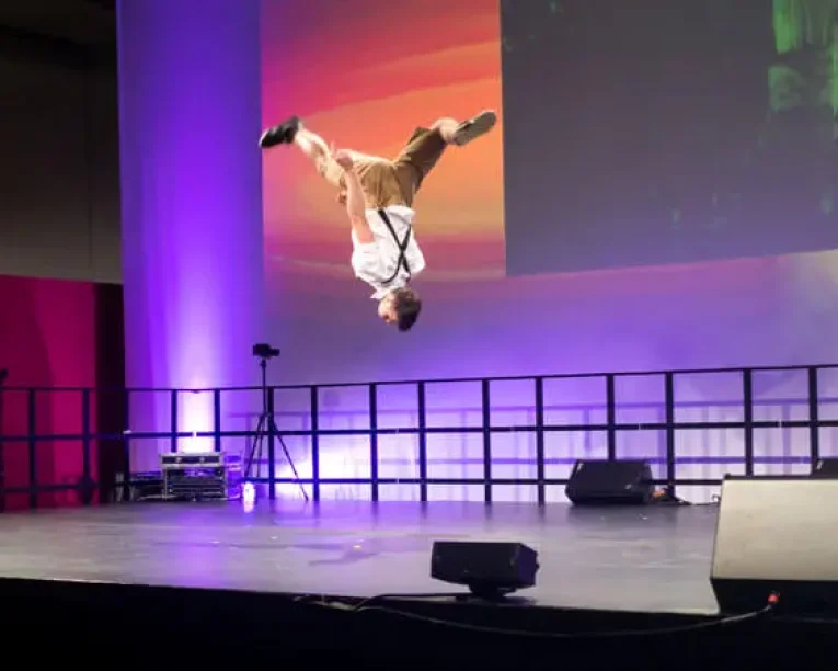 Ein Basketball-Freestyler macht in der Luft einen Rückwärtssalto auf der Bühne, mit lila Beleuchtung und einem bunten Hintergrund dahinter. Auf dem Boden sind Bühnengeräte und Lautsprecher zu sehen.