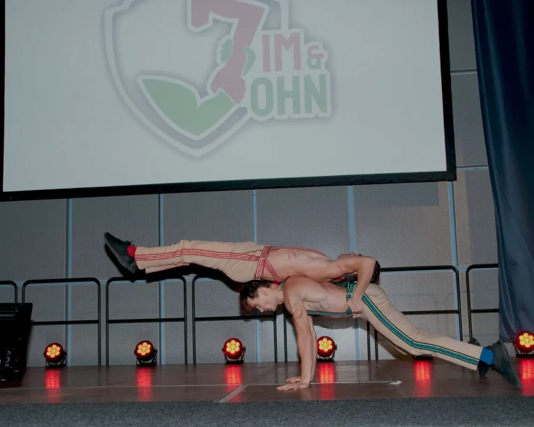 Zwei Akrobaten des Duo Comedy Akrobatik balancieren auf der Bühne; einer hält eine Planche, während der andere horizontal auf dem Rücken balanciert. Beide tragen gestreifte Hosen, hinter ihnen sind eine große Leinwand und Bühnenscheinwerfer zu sehen.