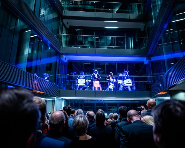 Eine Menschenmenge beobachtet eine Gruppe von Künstlern in beleuchteten Kostümen, die auf einem Balkon in einem modernen Glasgebäude Schlagzeug spielen, wobei blaue und lila Lichter eine futuristische Atmosphäre schaffen.